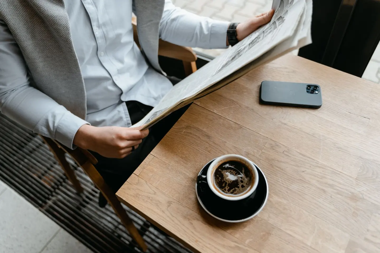 An adult male enjoying his coffee while reading a newspaper at a cafe with his smartphone nearby.
